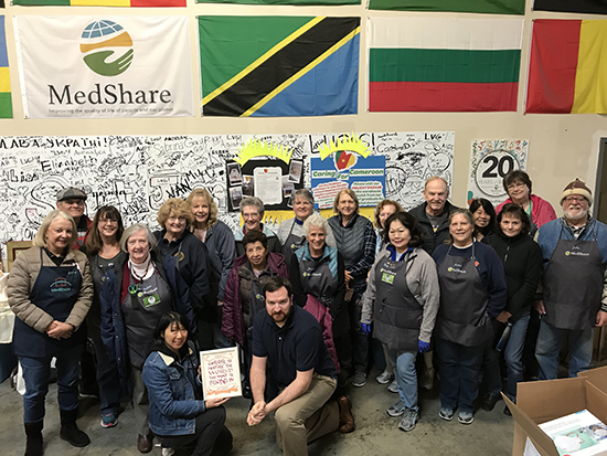 A group of about two dozen volunteers wearing aprons. They are in a warehouse. Behind them is a wall covered in different countries' flags. In front of them are the artist holding the certificate and Eric Talbert.